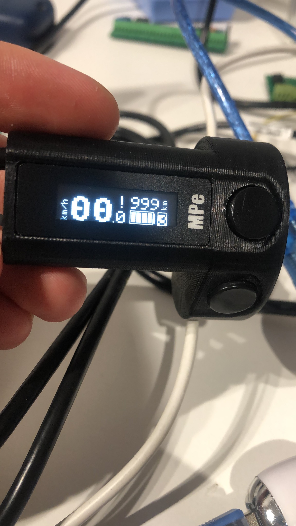 Czarny, trójwymiarowy wydrukowany miernik z wyświetlaczem LCD wskazującym prędkość i poziom naładowania baterii, trzymany w dłoni na tle kabli i elektroniki.