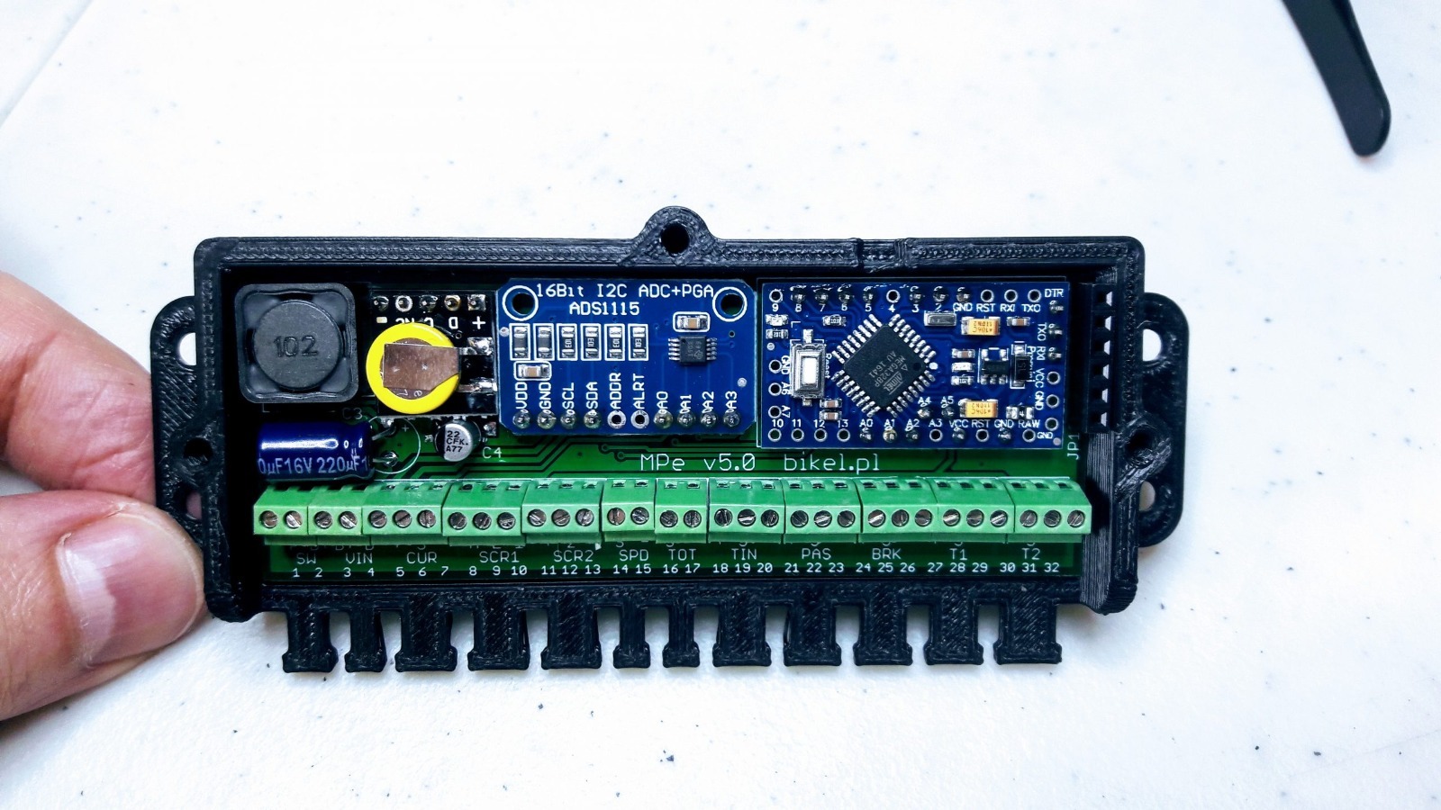 Elektroniczny moduł MPe v5.0 bikel.pl z elementami takimi jak 16Bit I2C ADC+PGA ADS1115, mikrokontroler, kondensatory i złącza śrubowe, umieszczony w czarnej obudowie drukowanej 3D, widziany z góry.