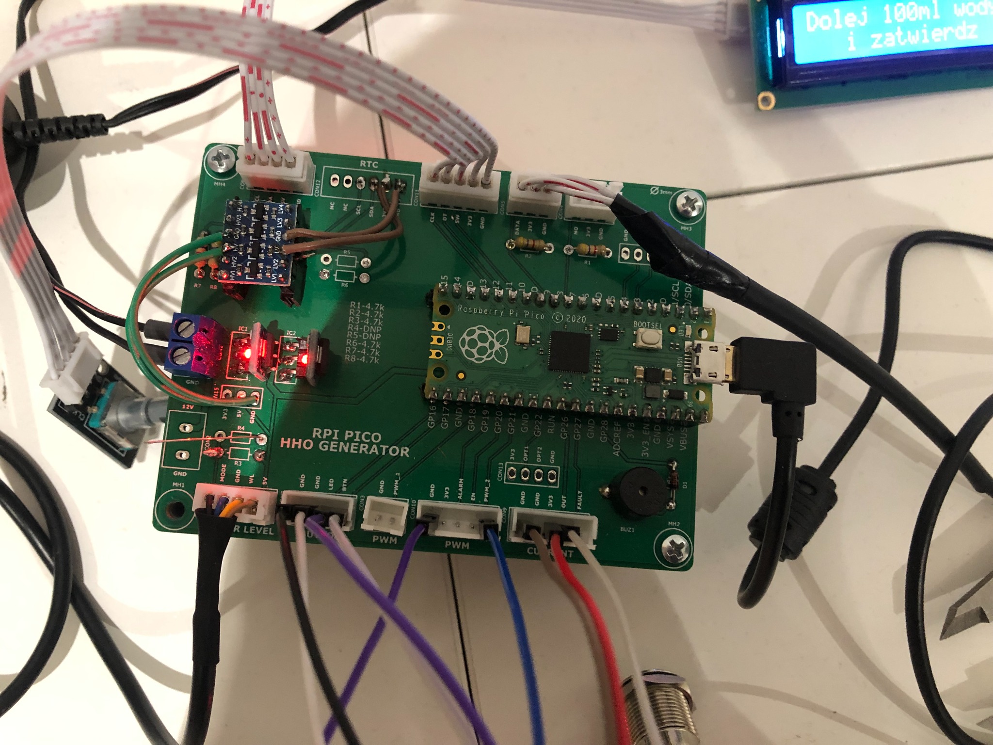 Zielona płytka RPI Pico HHO Generator z podłączonymi przewodami i modułami, widoczne elementy elektroniczne, rezystory, kondensatory i złącza, na białym tle.