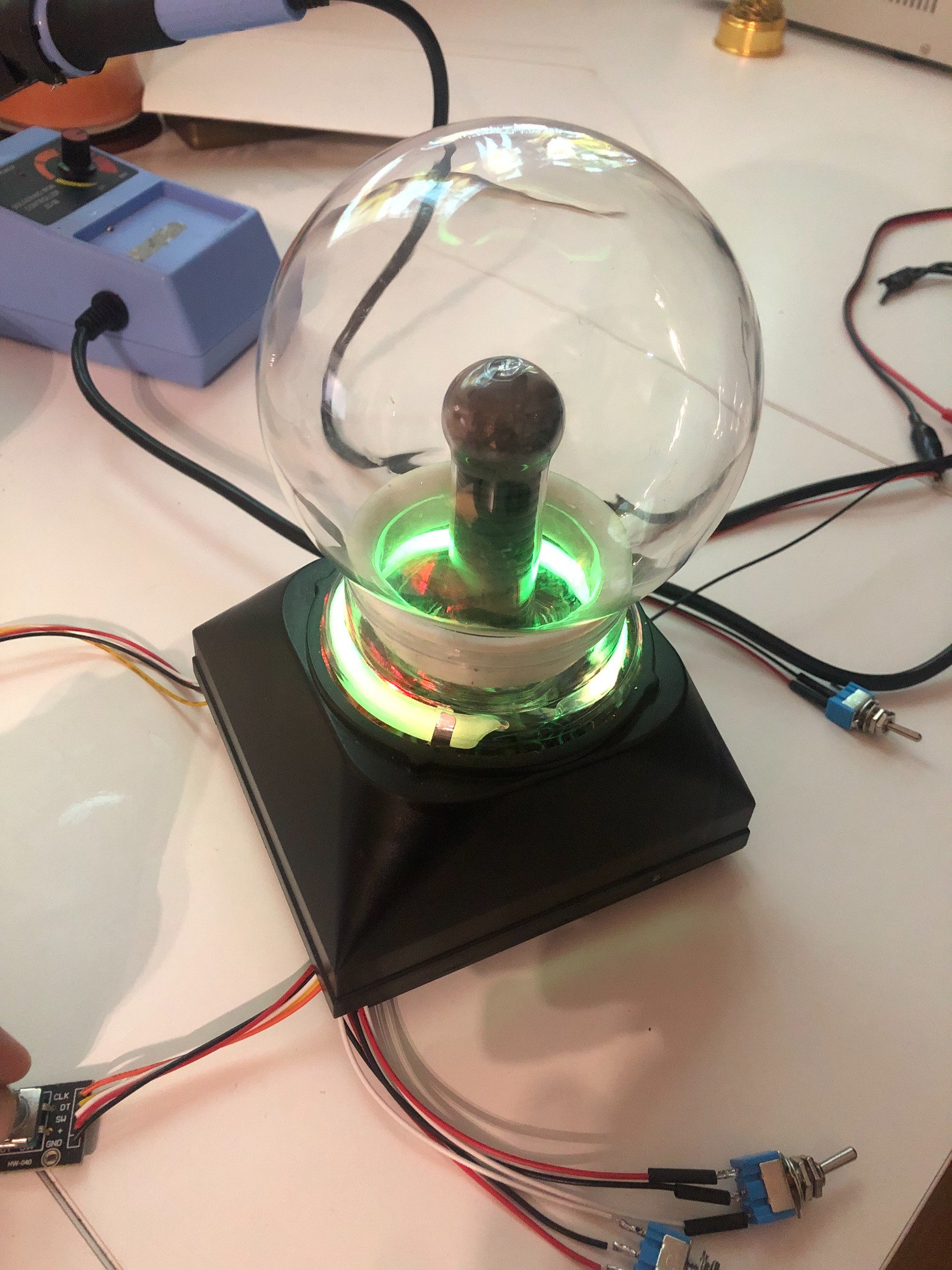 Szklana kula plazmowa z zielonym podświetleniem, widoczne podłączenia elektryczne i elementy lutownicze na białym stole.