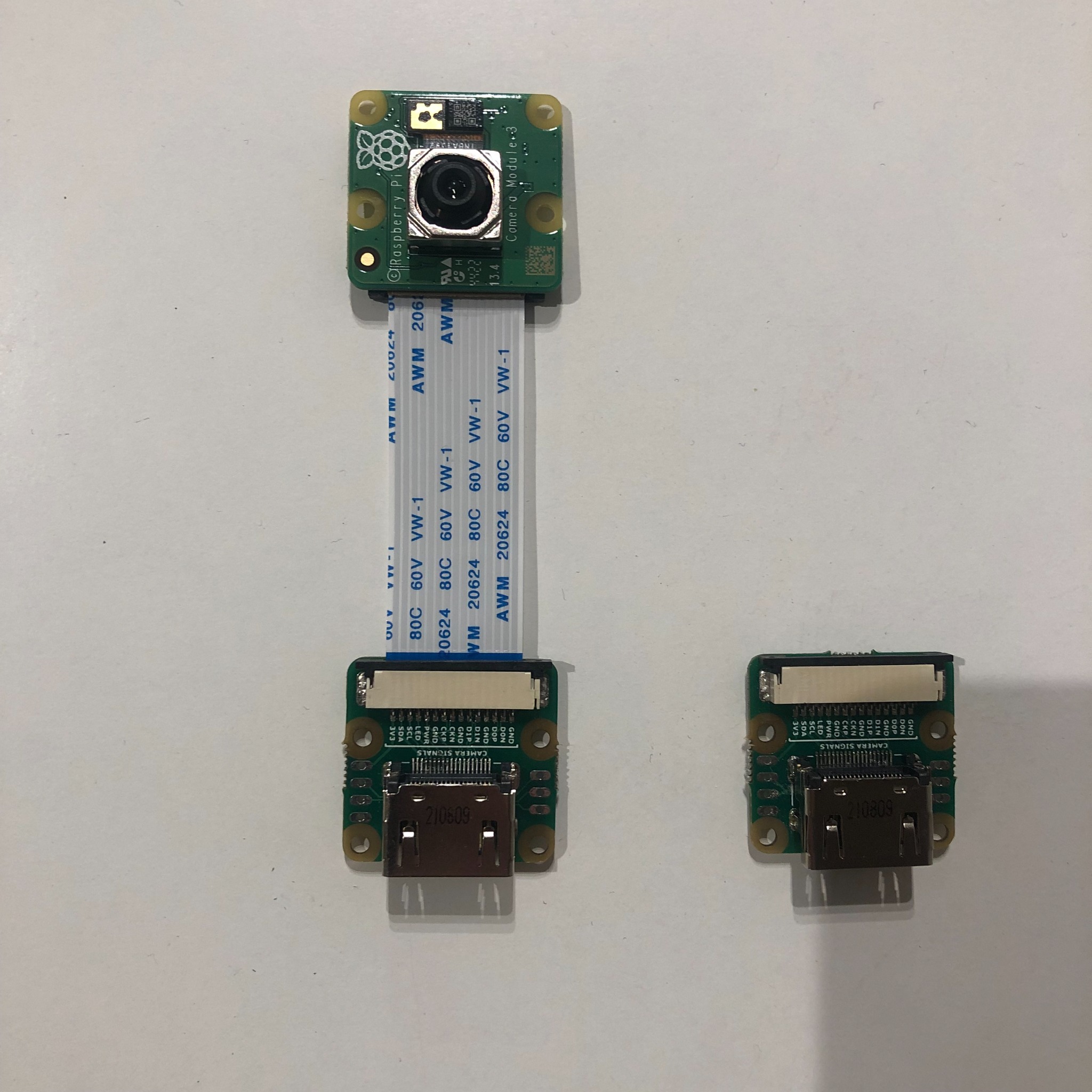 Raspberry Pi Camera Module V3, z kablem taśmowym i dwoma złączami, leżące na białym tle. Widoczne detale elektroniki i oznaczenia na płytkach.
