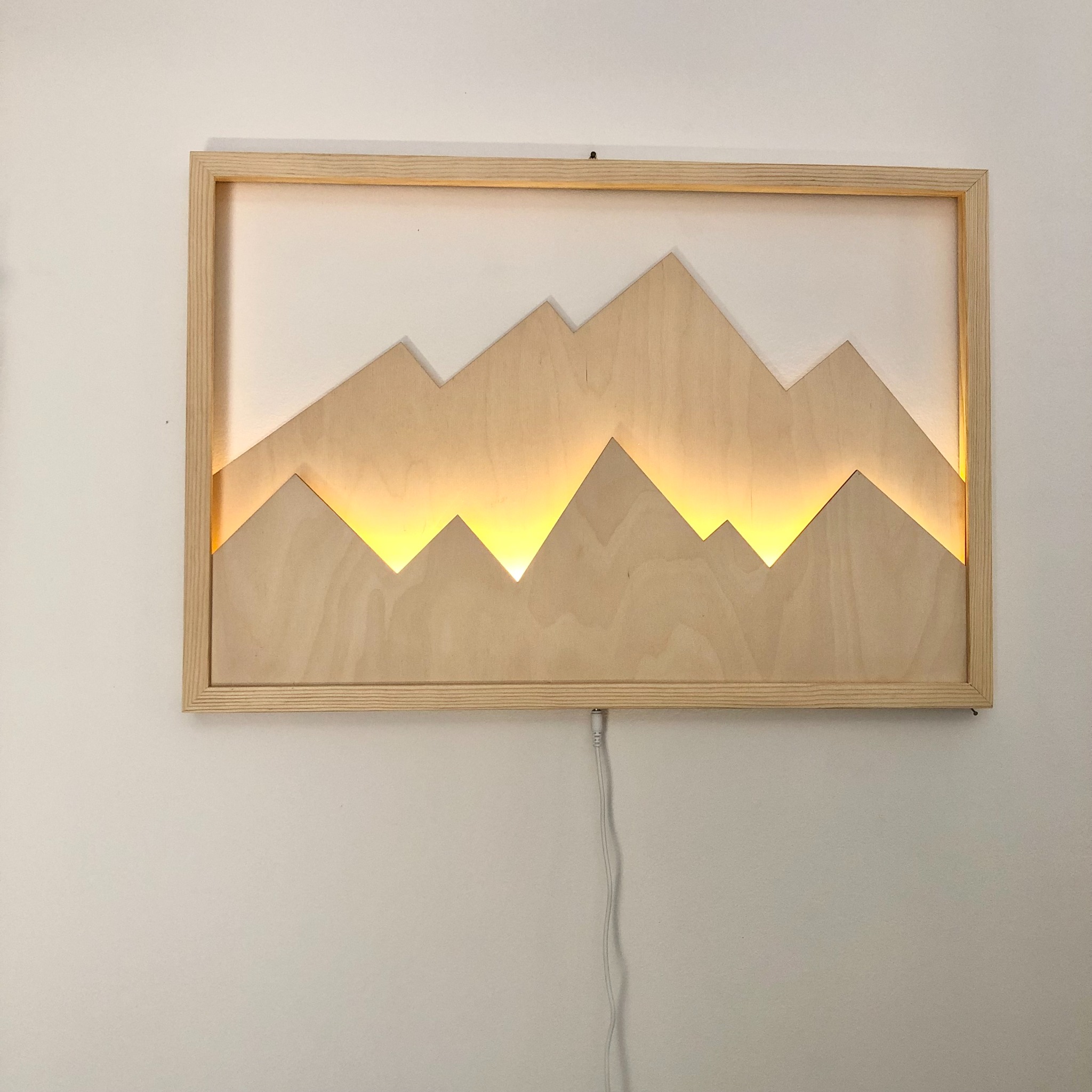 Drewniana, podświetlana dekoracja ścienna w kształcie gór w jasnej ramie, zawieszona na białej ścianie, z widocznym kablem zasilającym.