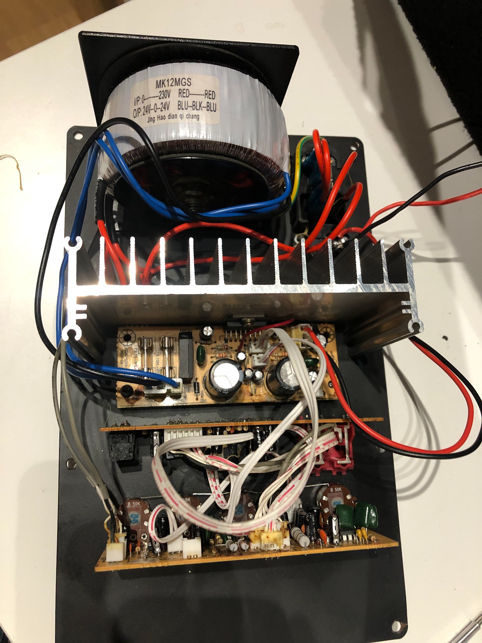 Wnętrze urządzenia elektronicznego z widocznymi płytkami drukowanymi, transformatorem z etykietą MK12MGS, radiatorem i licznymi przewodami w kolorach czerwonym, niebieskim i czarnym.