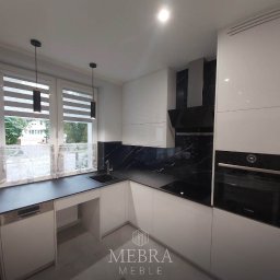 Rafał Rytwiński - Nowoczesna kuchnia z białymi szafkami, czarnym blatem i marmurowym backsplashiem, widok na okno z roletami.