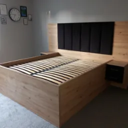 Łóżko z drewnianą ramą i czarnym, tapicerowanym zagłówkiem, z wbudowanymi szafkami nocnymi z szufladami, na tle ściany z dekoracjami.