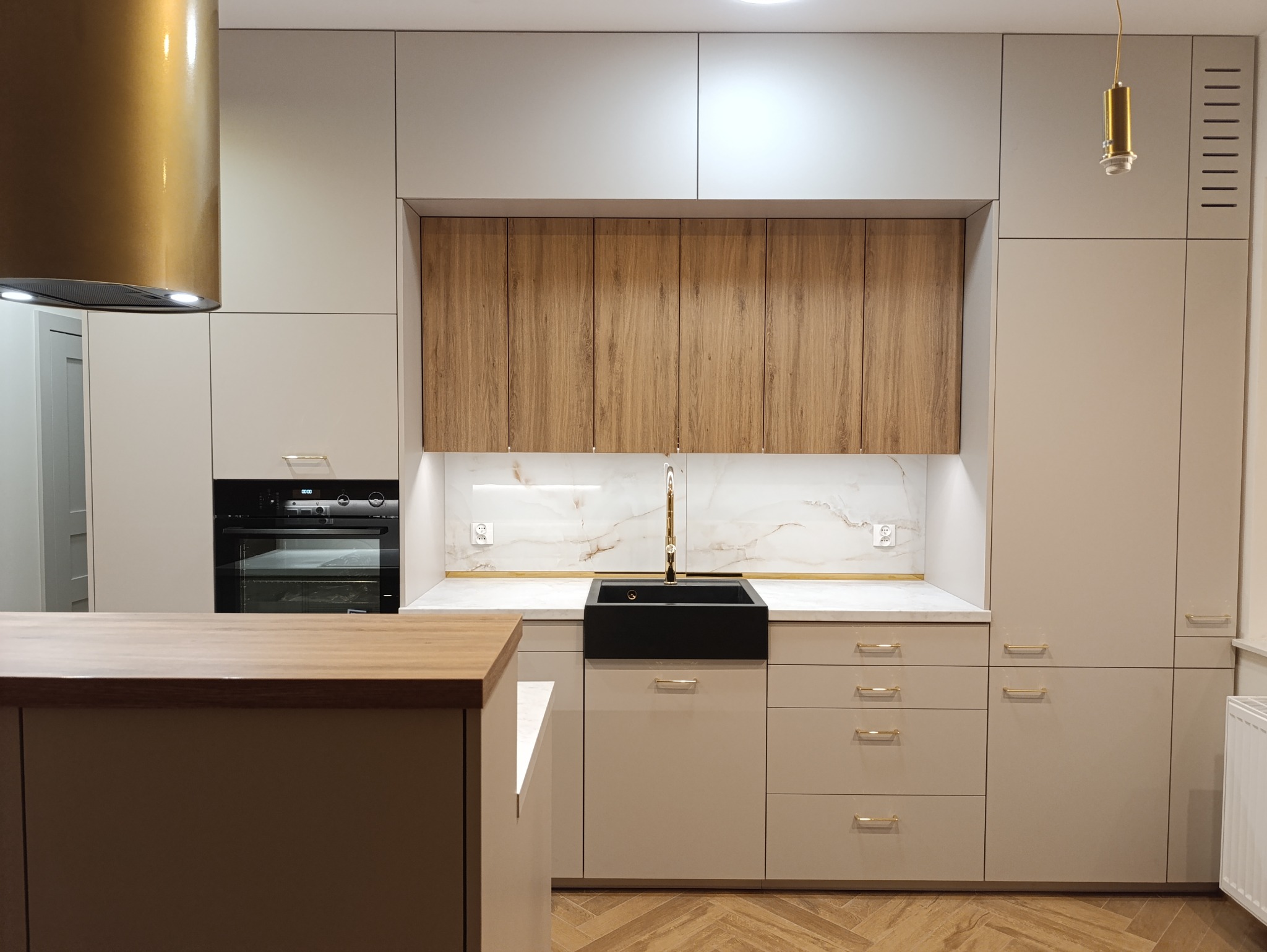 Nowoczesna kuchnia w odcieniach beżu i drewna z czarnym zlewem farmerskim i złotą armaturą, parkiet w jodełkę na podłodze.