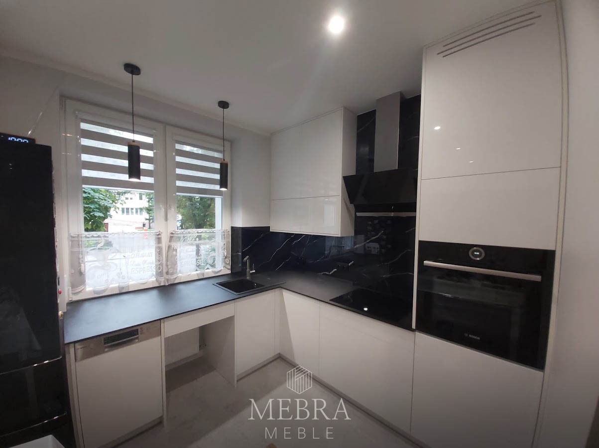 Nowoczesna kuchnia z białymi szafkami, czarnym blatem i marmurowym backsplashiem, widok na okno z roletami.