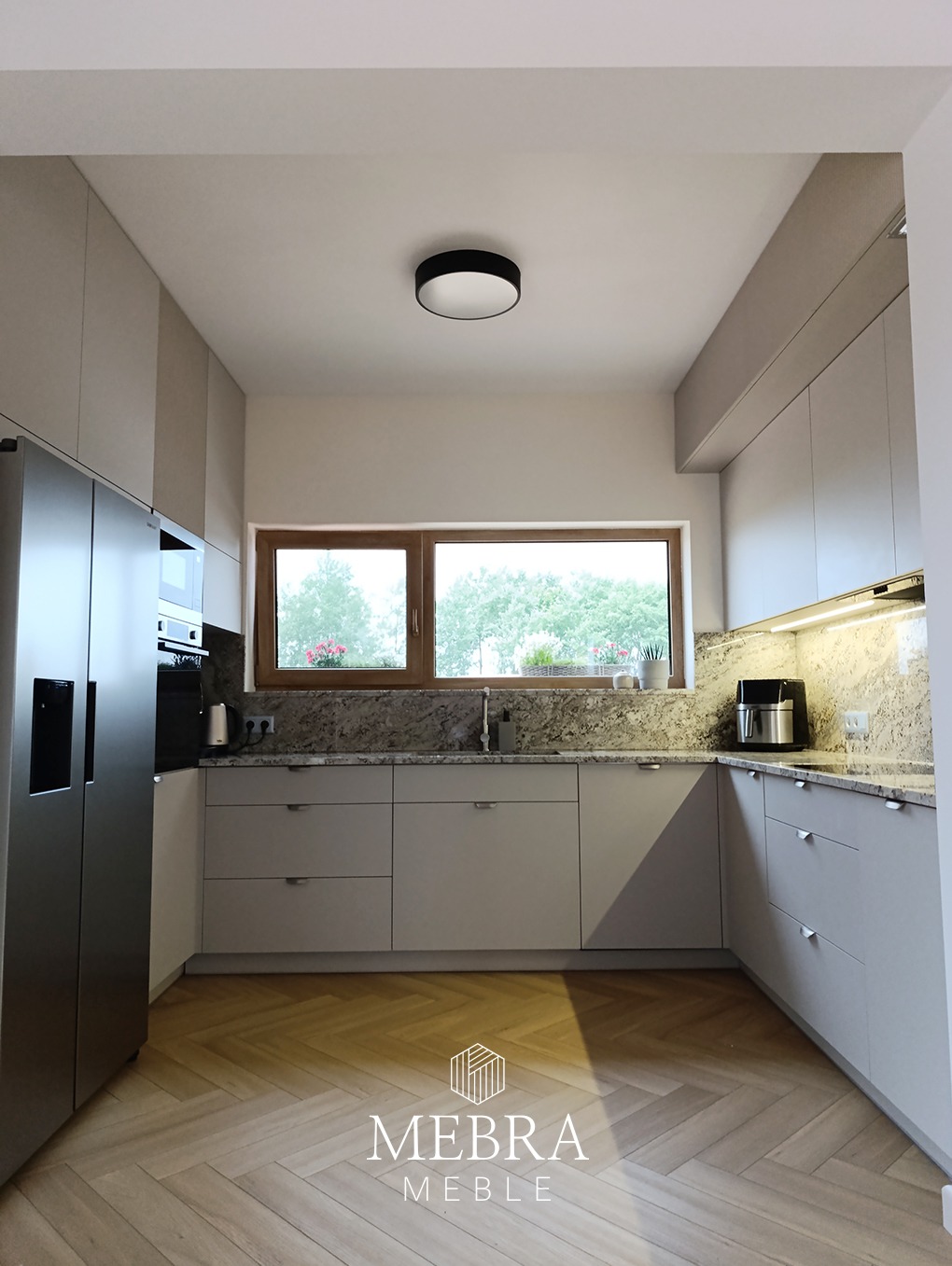 Jasna kuchnia z szarymi, matowymi frontami szafek, drewnianym blatem i podłogą ułożoną w jodełkę, z widocznym logo firmy na pierwszym planie.