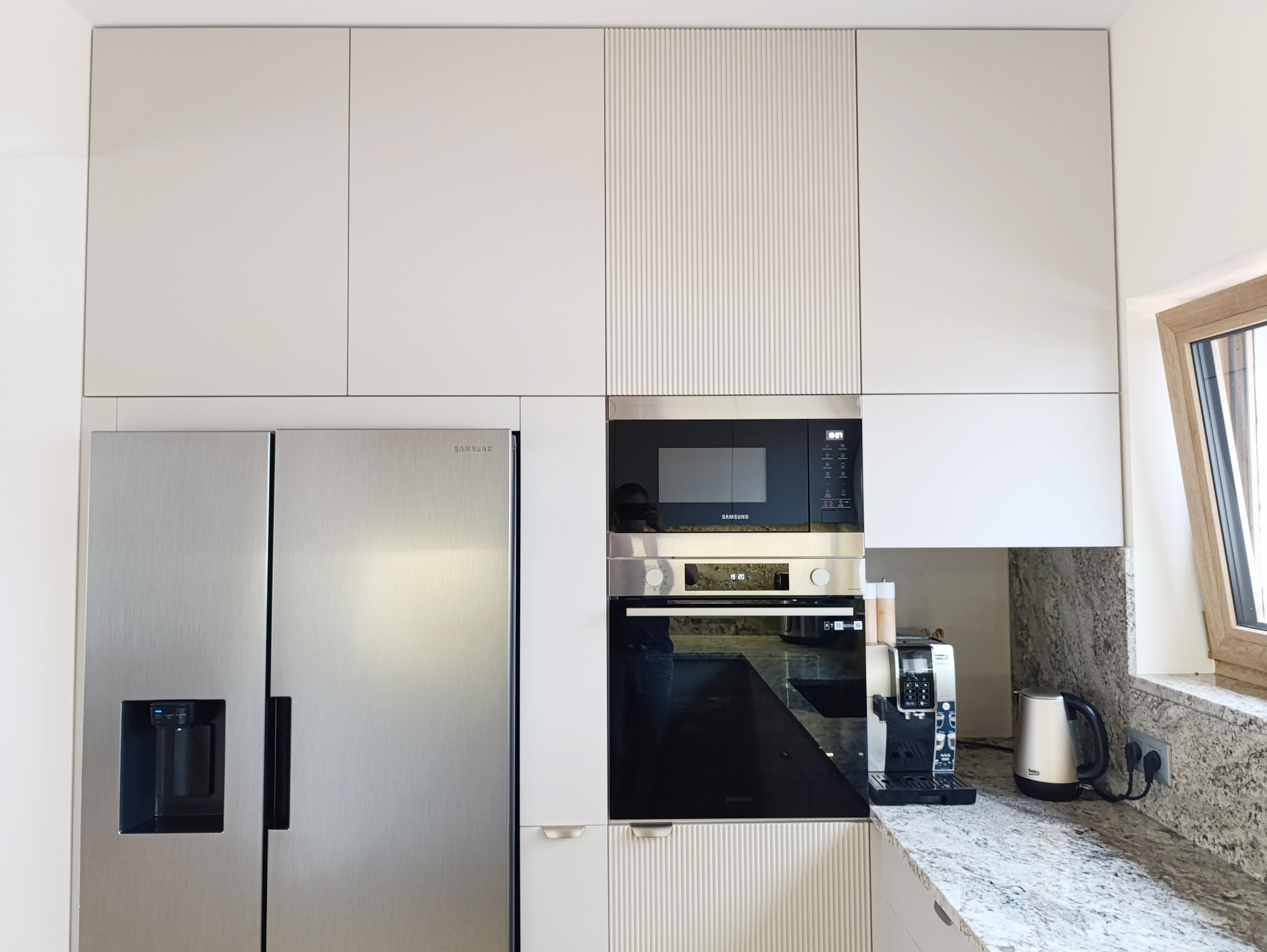 Nowoczesna, jasna kuchnia z zabudową meblową: srebrna lodówka side-by-side, piekarnik i mikrofalówka w zabudowie, białe szafki górne z elementami ryflowanymi, blat z jasnego granitu, ekspres...