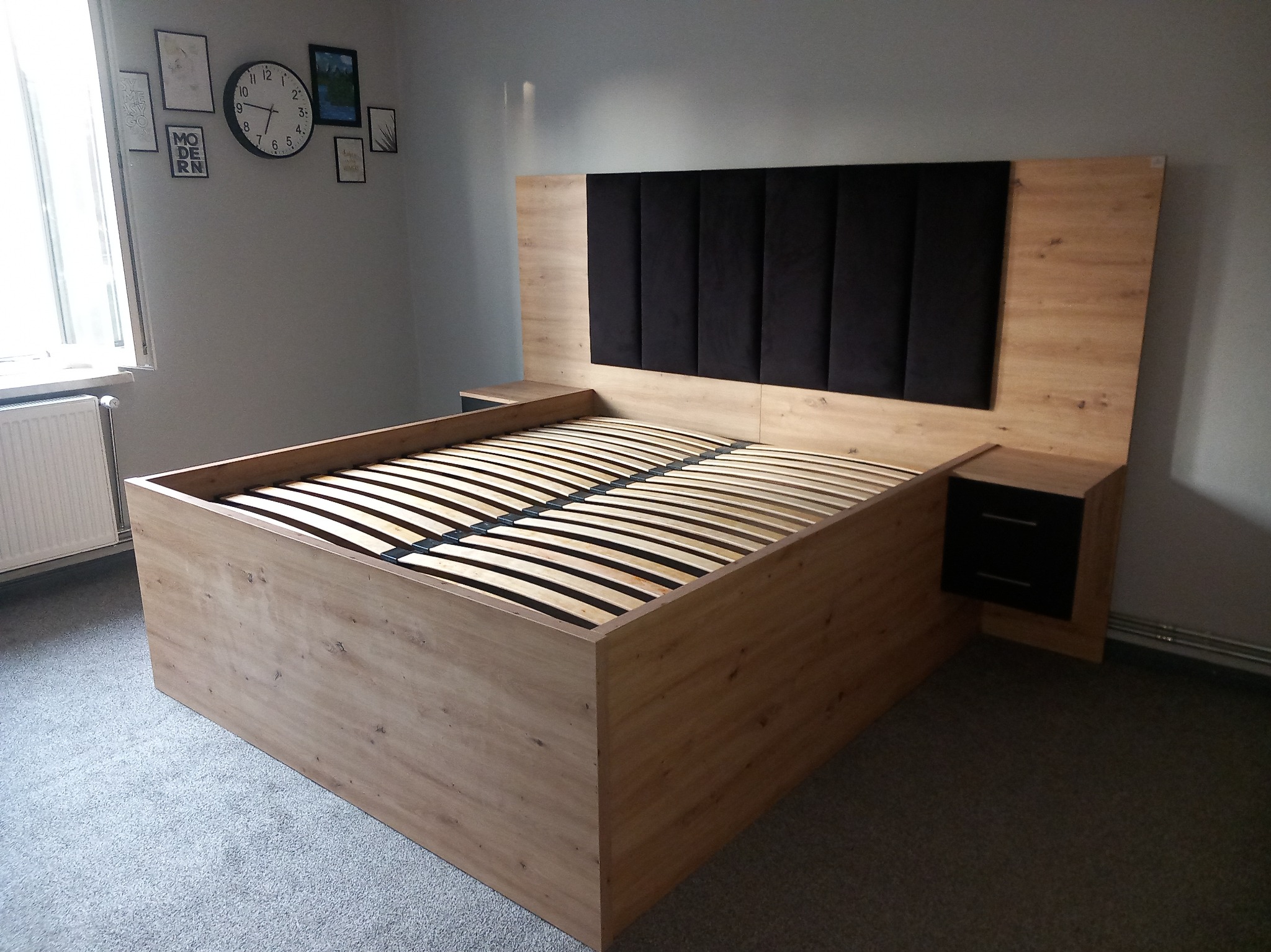 Łóżko z drewnianą ramą i czarnym, tapicerowanym zagłówkiem, z wbudowanymi szafkami nocnymi z szufladami, na tle ściany z dekoracjami.