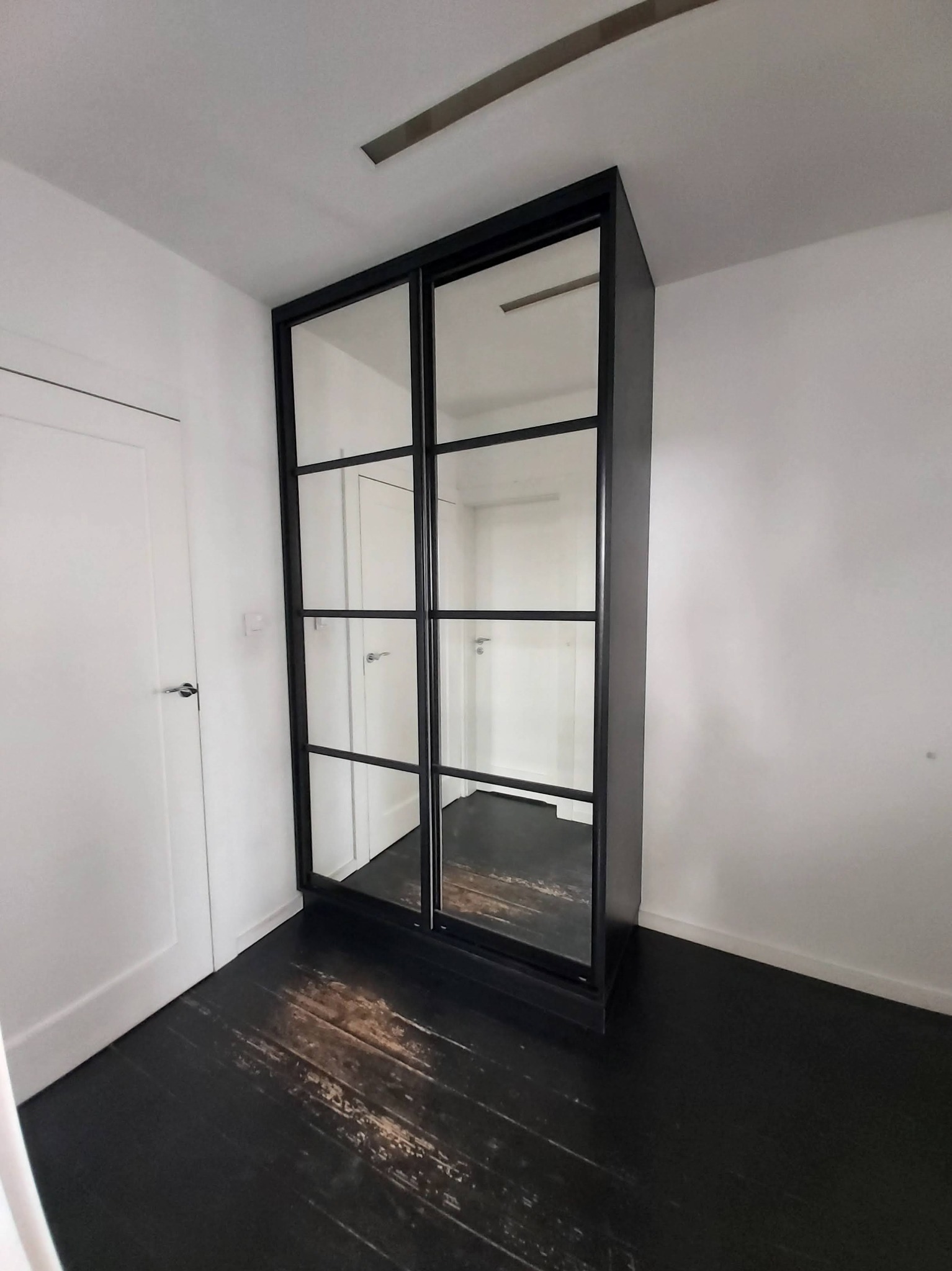Czarna szafa wnękowa z przesuwanymi drzwiami z lustrami w ramie, widoczna na tle białych ścian i ciemnej, drewnianej podłogi.