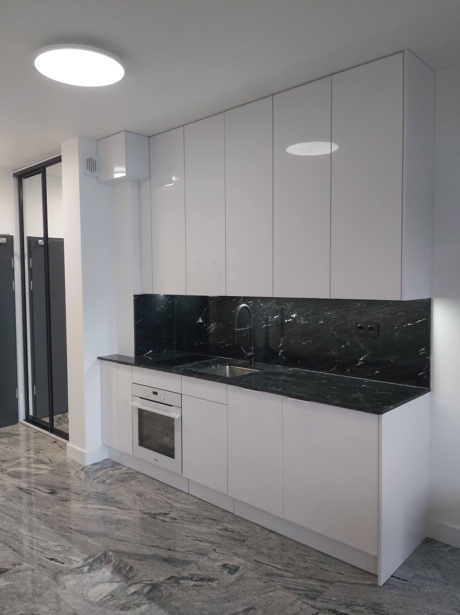 Nowoczesna kuchnia z białymi, lakierowanymi szafkami, czarnym blatem z marmurowym wzorem i szarą marmurową podłogą, oświetlona górnym światłem.