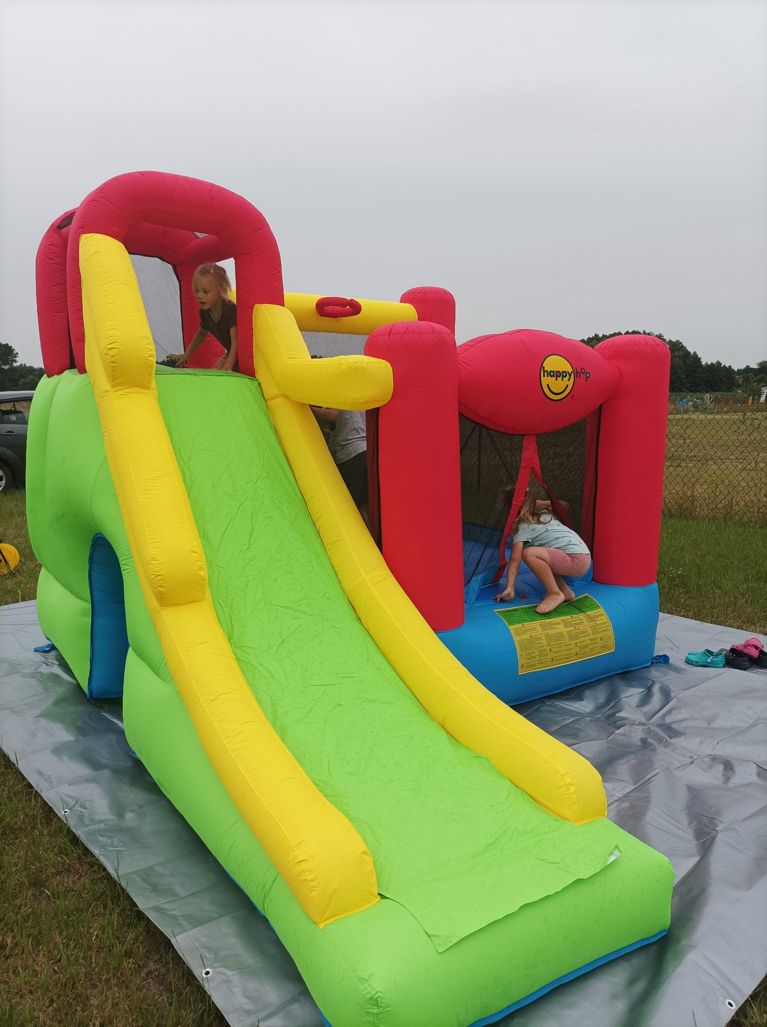Dzieci bawią się na kolorowym dmuchanym zamku ze zjeżdżalnią na trawniku, z widocznym logo 'happy hop' na czerwonej ściance.