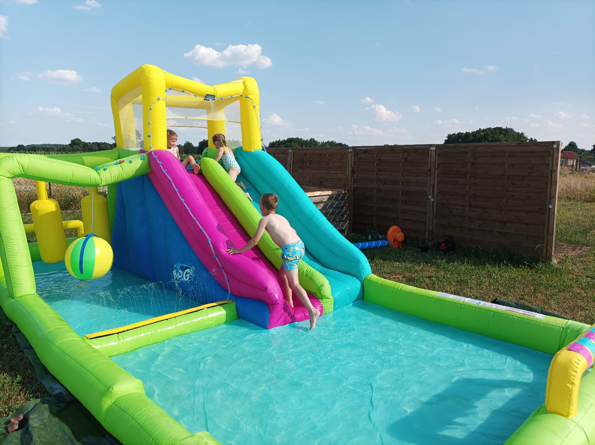 Dzieci bawią się na kolorowym, dmuchanym placu zabaw z basenem i zjeżdżalniami na trawniku w słoneczny dzień.
