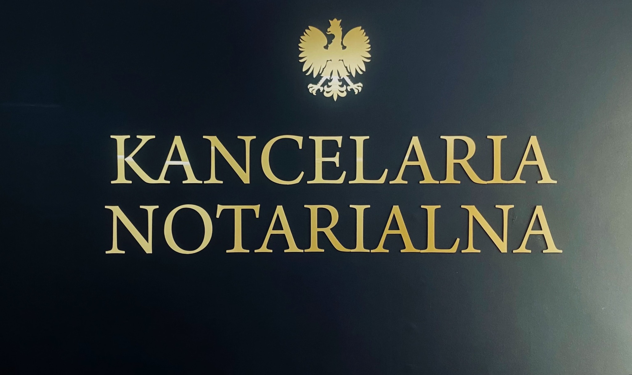 Złoty napis 'Kancelaria Notarialna' oraz godło Polski na ciemnym tle ściany.
