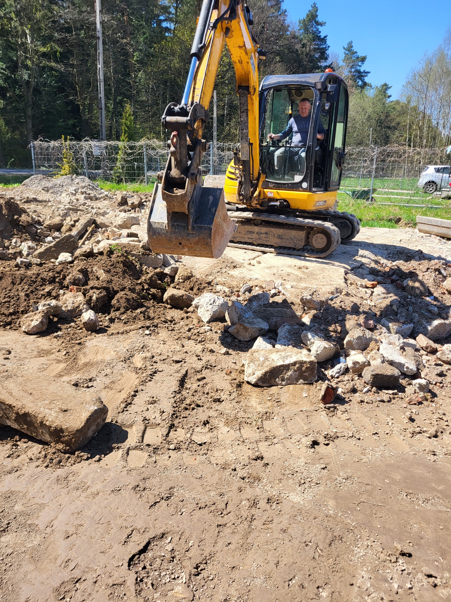 Żółta minikoparka z operatorem w kabinie, pracująca na placu budowy, gdzie widoczne są gruz, ziemia i duże kamienie, z lasem w tle pod błękitnym niebem.