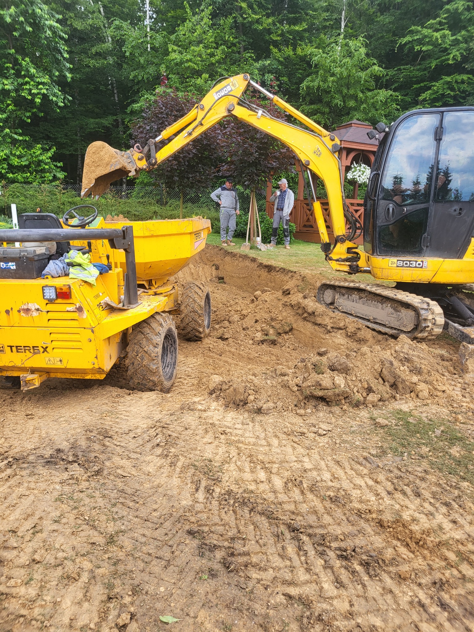 Żółta koparka nabiera ziemię do żółtej wywrotki; w tle dwóch mężczyzn stoi obok wykopu w ziemi, za nimi altanka z kwiatami.