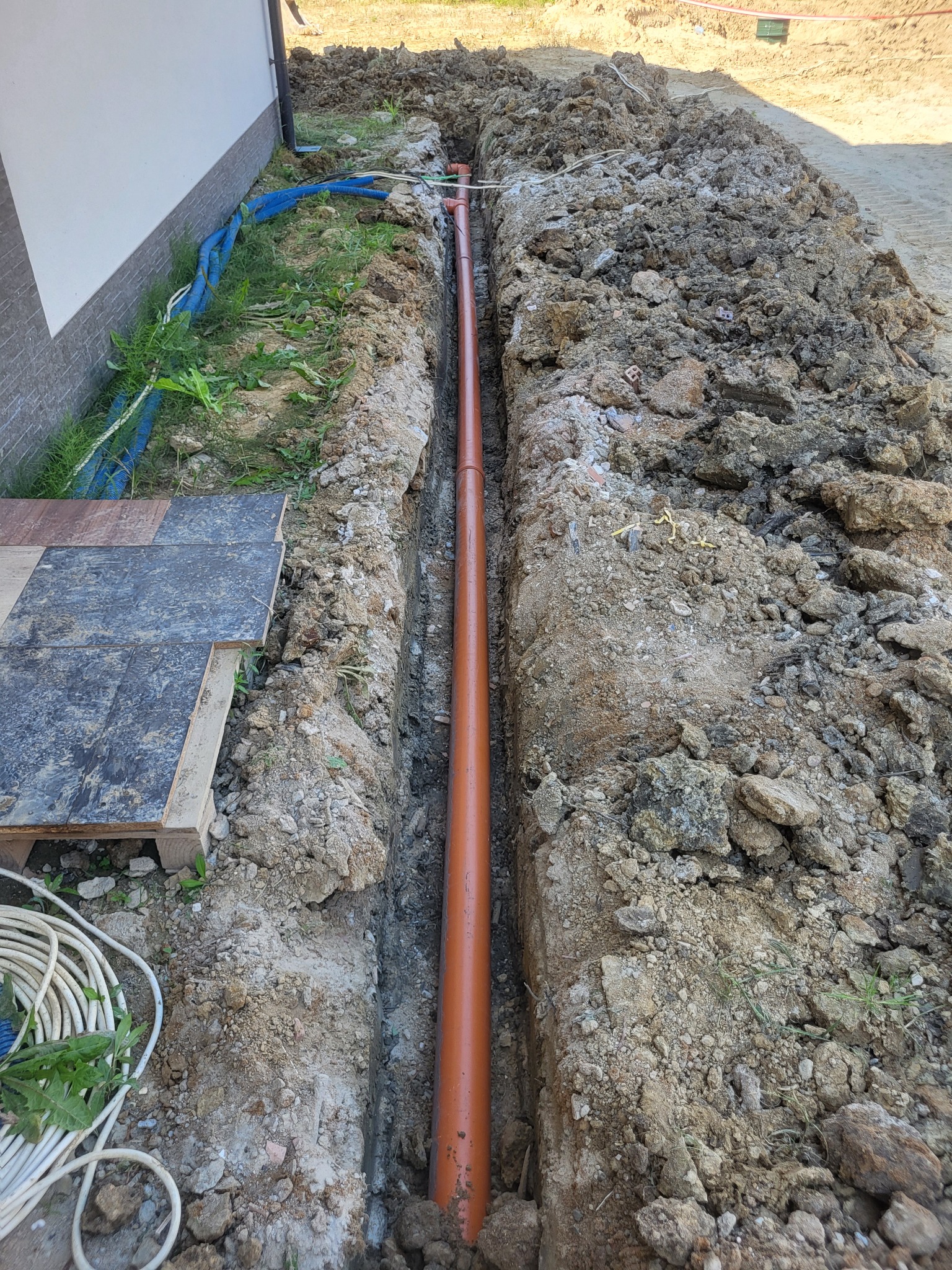 Wąski wykop pod rurę w Minodze, widoczne ułożone rury z tworzywa sztucznego i ziemia po bokach, fragment budynku w tle.