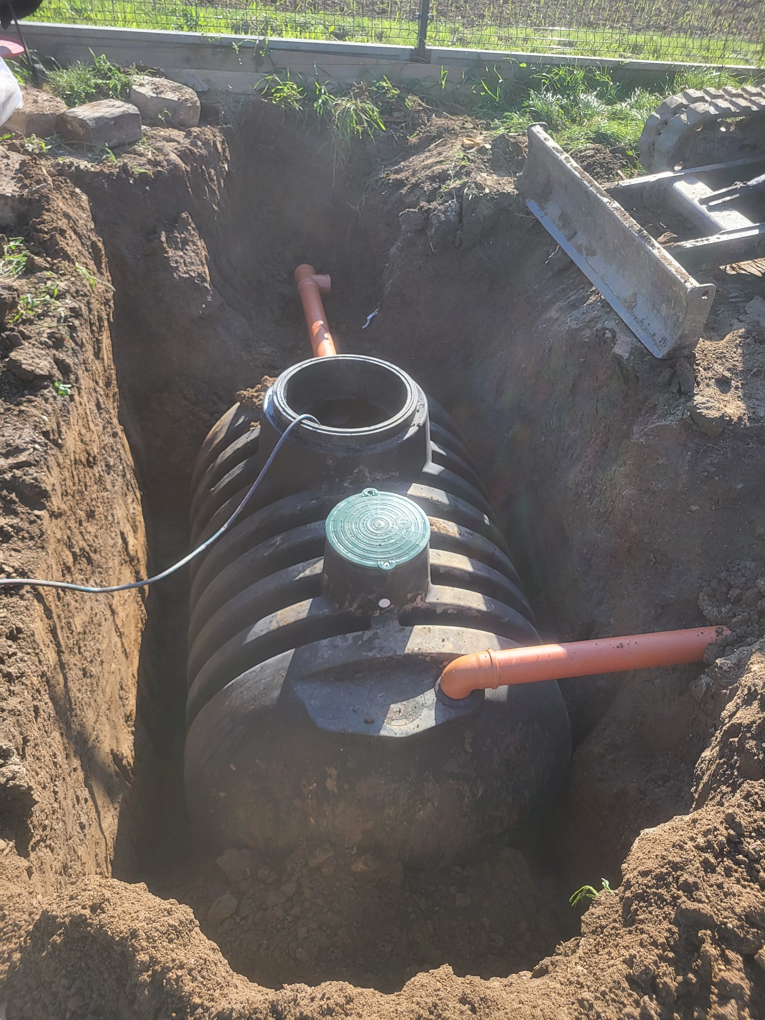 Czarny zbiornik z zieloną pokrywą umieszczony w głębokim wykopie z rurami przyłączeniowymi, widok z góry na dół.