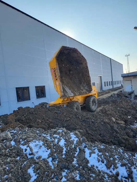 Żółta wywrotka Terex z uniesioną skrzynią, wysypująca ziemię na placu budowy, widoczne bryły ziemi z resztkami śniegu, w tle szary budynek przemysłowy z oknami.