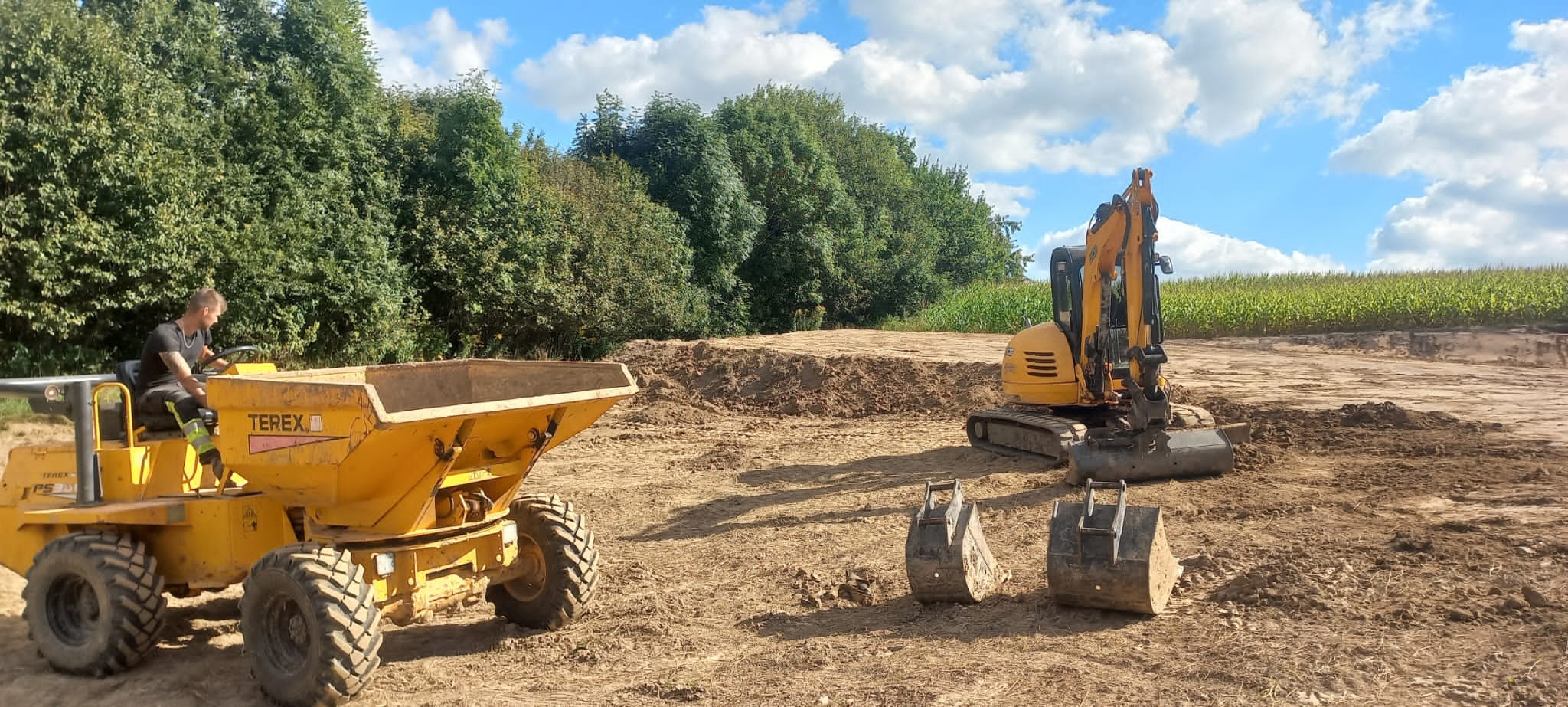 Żółta wywrotka Terex z operatorem i żółta koparka na gąsienicach na placu budowy z dwoma łyżkami obok, w tle drzewa i pole uprawne pod błękitnym niebem z chmurami.