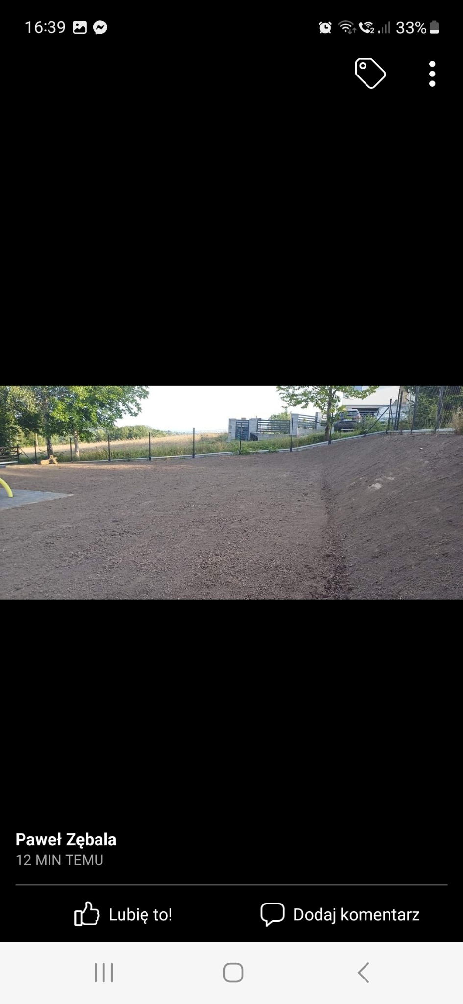 Równy teren pokryty ciemną ziemią, ogrodzony metalową siatką, z widocznymi drzewami i budynkiem w tle.