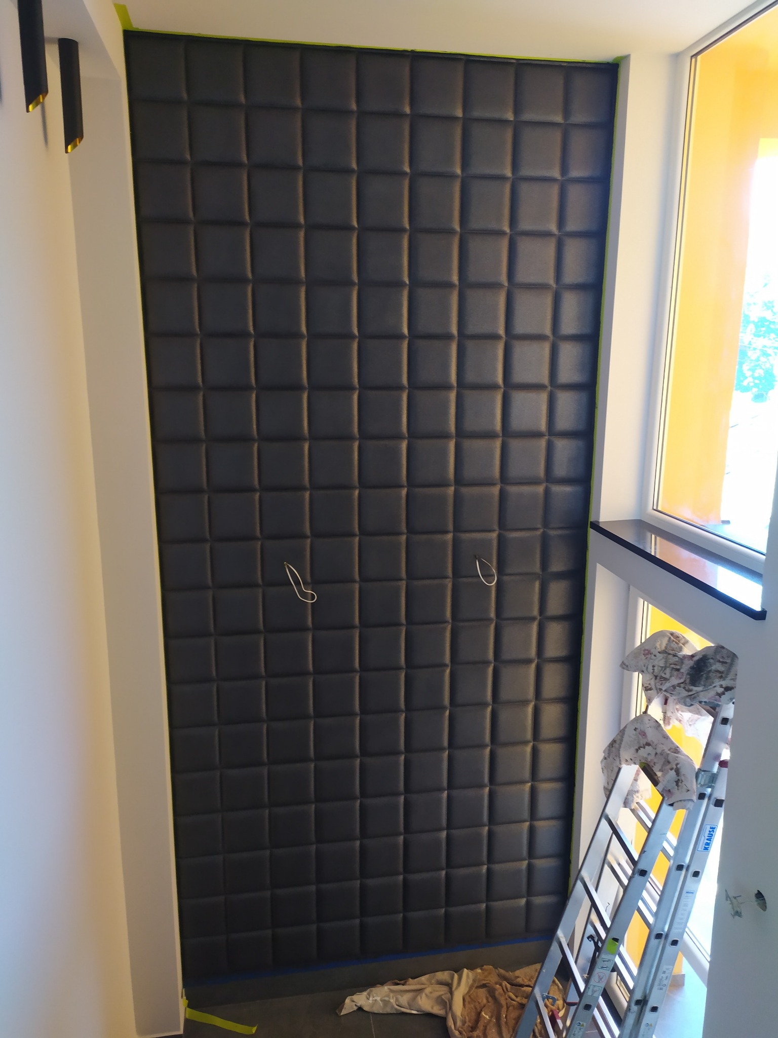 Ciemnoszara, pikowana ściana z paneli, zabezpieczona taśmą malarską przy suficie, widoczna drabina i akcesoria malarskie w narożniku, okno z żółtą folią ochronną.