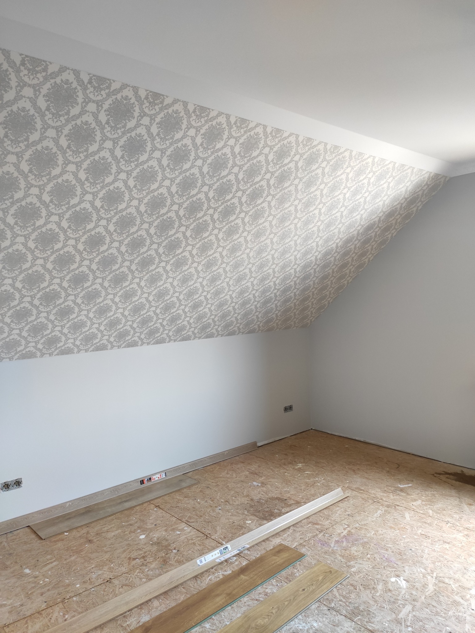 Pokój na poddaszu w trakcie prac: ściana z szarą tapetą w ornamenty na skosie, biała ściana boczna, surowa podłoga z płyt OSB z przygotowanymi panelami podłogowymi.