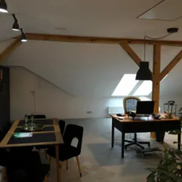 Biuro w loftowym stylu z drewnianymi belkami stropowymi, biurkiem z laptopem, stołem konferencyjnym i roślinami doniczkowymi, doświetlone naturalnym światłem z okna dachowego.