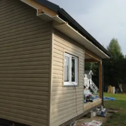 Boczny widok domu z drewnianą elewacją w trakcie montażu, widoczne elementy konstrukcyjne i narzędzia na trawniku.