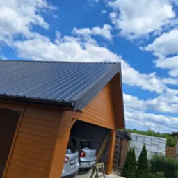 Nowoczesny garaż z brązową drewnianą elewacją i ciemnoszarym dachem z blachy trapezowej na tle błękitnego nieba z białymi chmurami, obok budynek w stanie surowym.