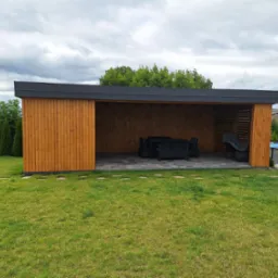 Nowoczesna drewniana altana z płaskim dachem, wyposażona w czarne meble ogrodowe, usytuowana na trawniku z widokiem na basen w tle.