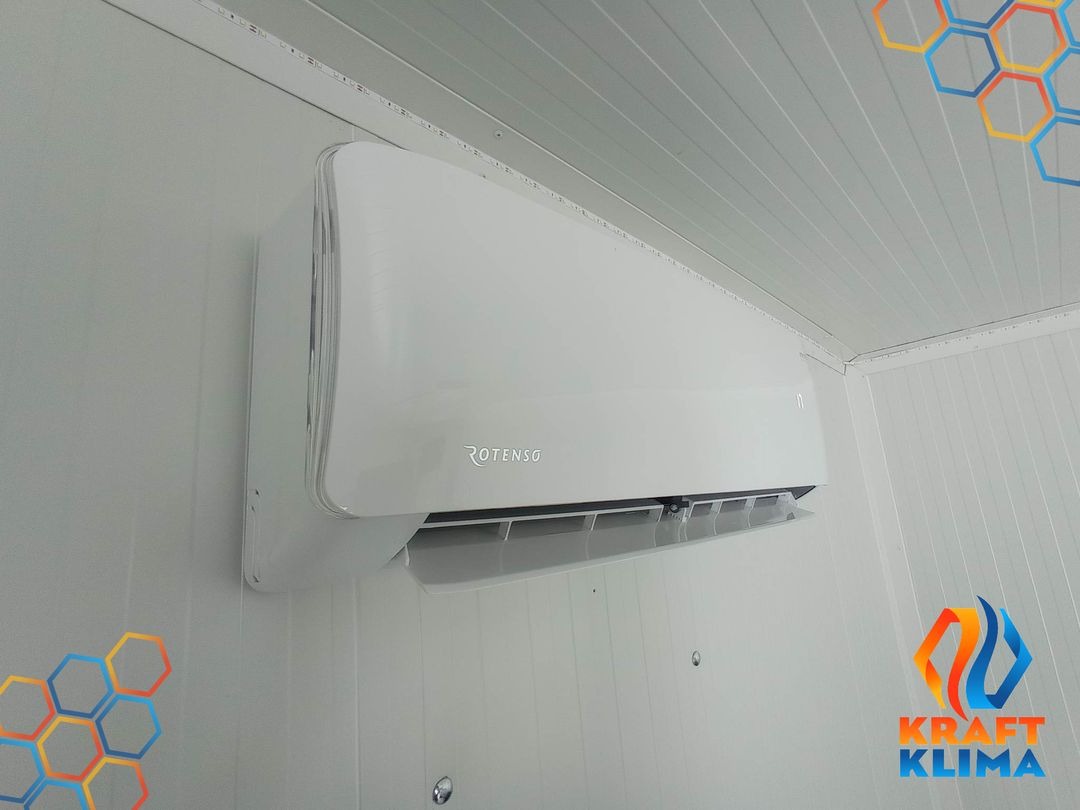 Biały klimatyzator Rotenso zamontowany na ścianie wyłożonej panelami PCV, widoczne kolorowe naklejki o strukturze plastra miodu na ścianie i suficie.