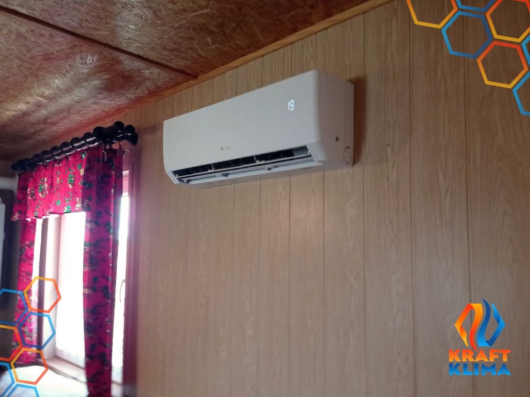 Biały klimatyzator z wyświetlaczem temperatury (19 stopni) zamontowany na ścianie z boazerii, widoczne zasłony w kwiaty i logo firmy w rogu zdjęcia.