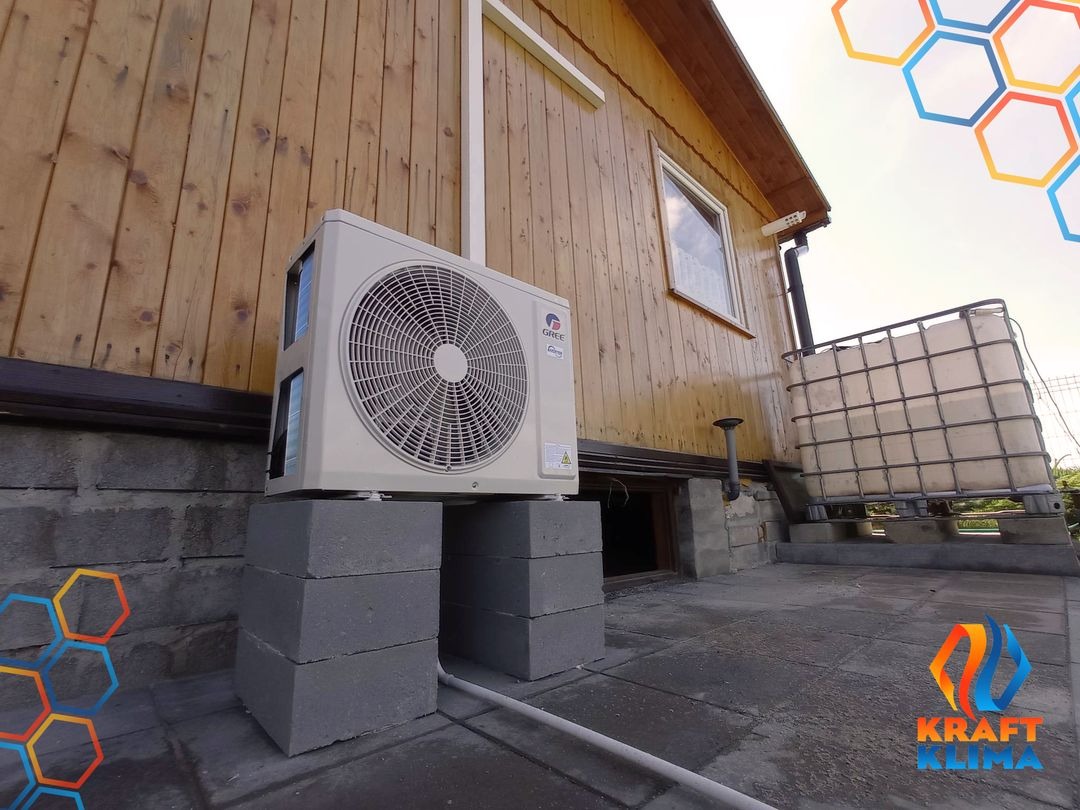 Zewnętrzna jednostka klimatyzatora marki Gree zamontowana na betonowych bloczkach przy drewnianej elewacji budynku jednorodzinnego, widoczny zbiornik na wodę i okno piwniczne.