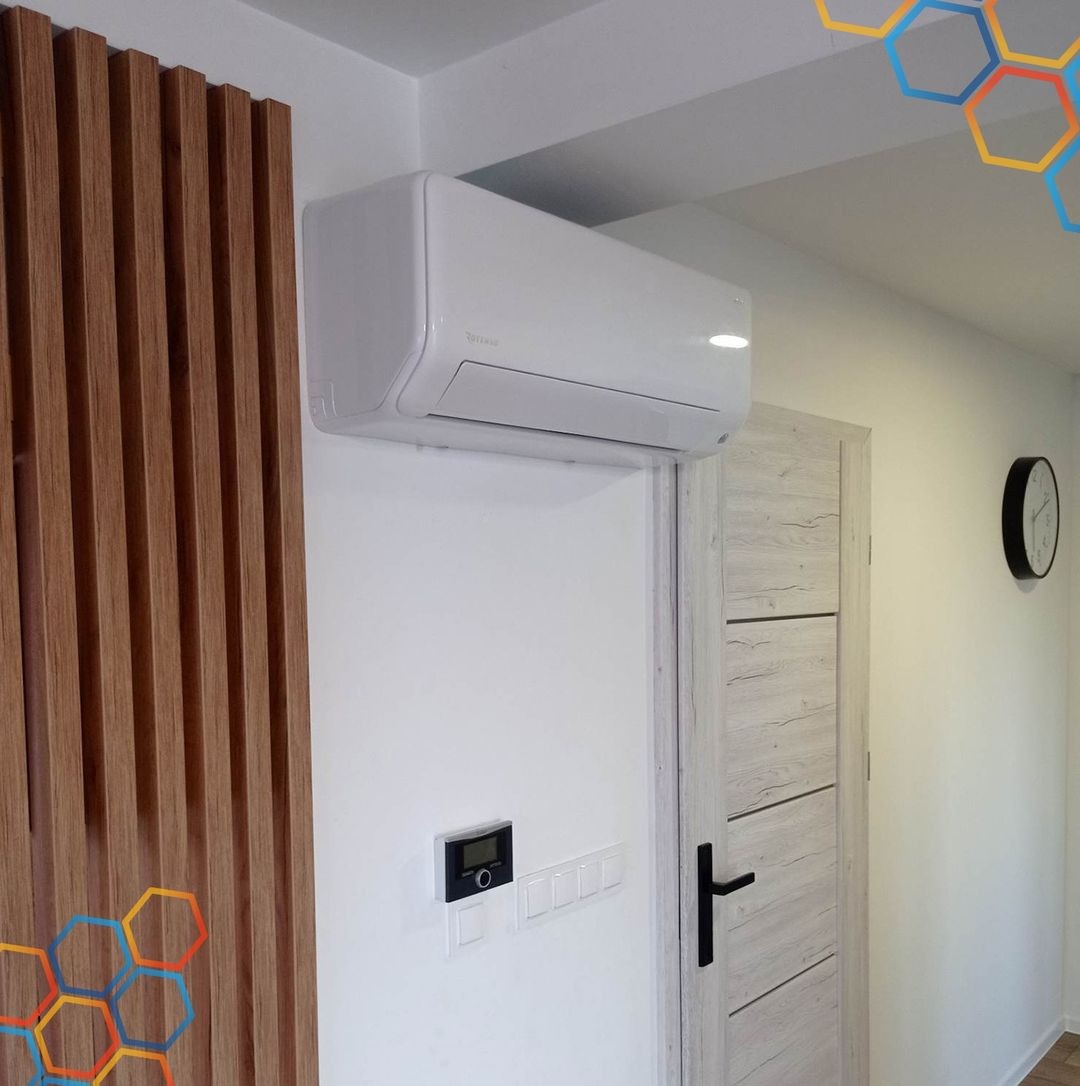 Biały klimatyzator ścienny zamontowany w nowoczesnym wnętrzu, widoczny panel sterowania i dekoracyjna drewniana ścianka lamelowa obok drzwi z jasnego drewna z czarną klamką.