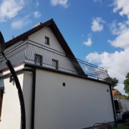 Biały budynek z balkonem zabezpieczonym czarną, ozdobną konstrukcją z metalu, na tle błękitnego nieba z chmurami.