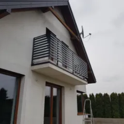 Nowoczesny balkon z czarną, poziomą barierką, widok z dołu na tle szarej elewacji domu w trakcie wykończenia i drabiny oparta o ścianę.