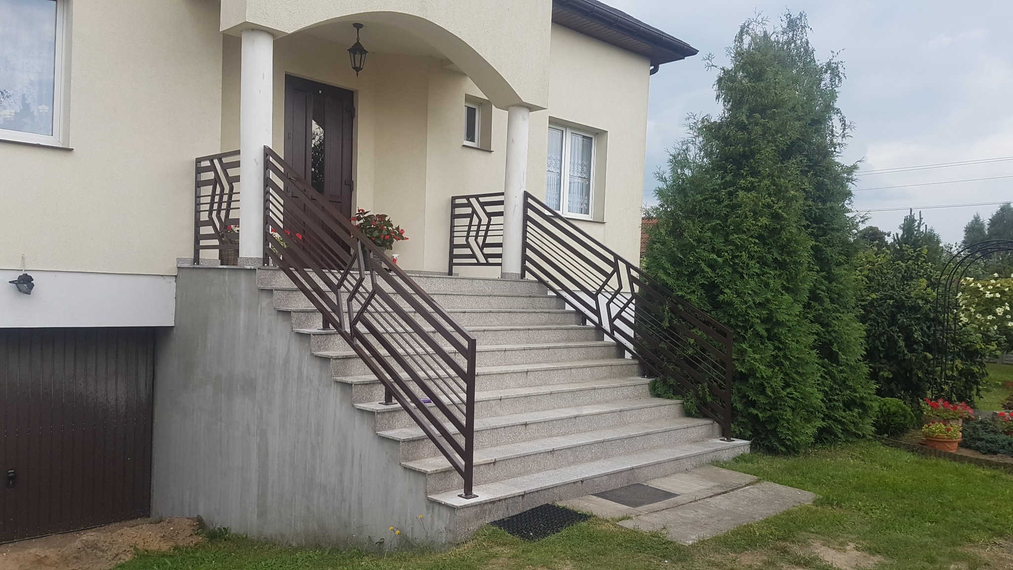 Zewnętrzne schody wyłożone jasnymi płytkami z brązową, metalową barierką o nowoczesnym wzorze, z donicami z kwiatami po bokach, prowadzące do wejścia domu.