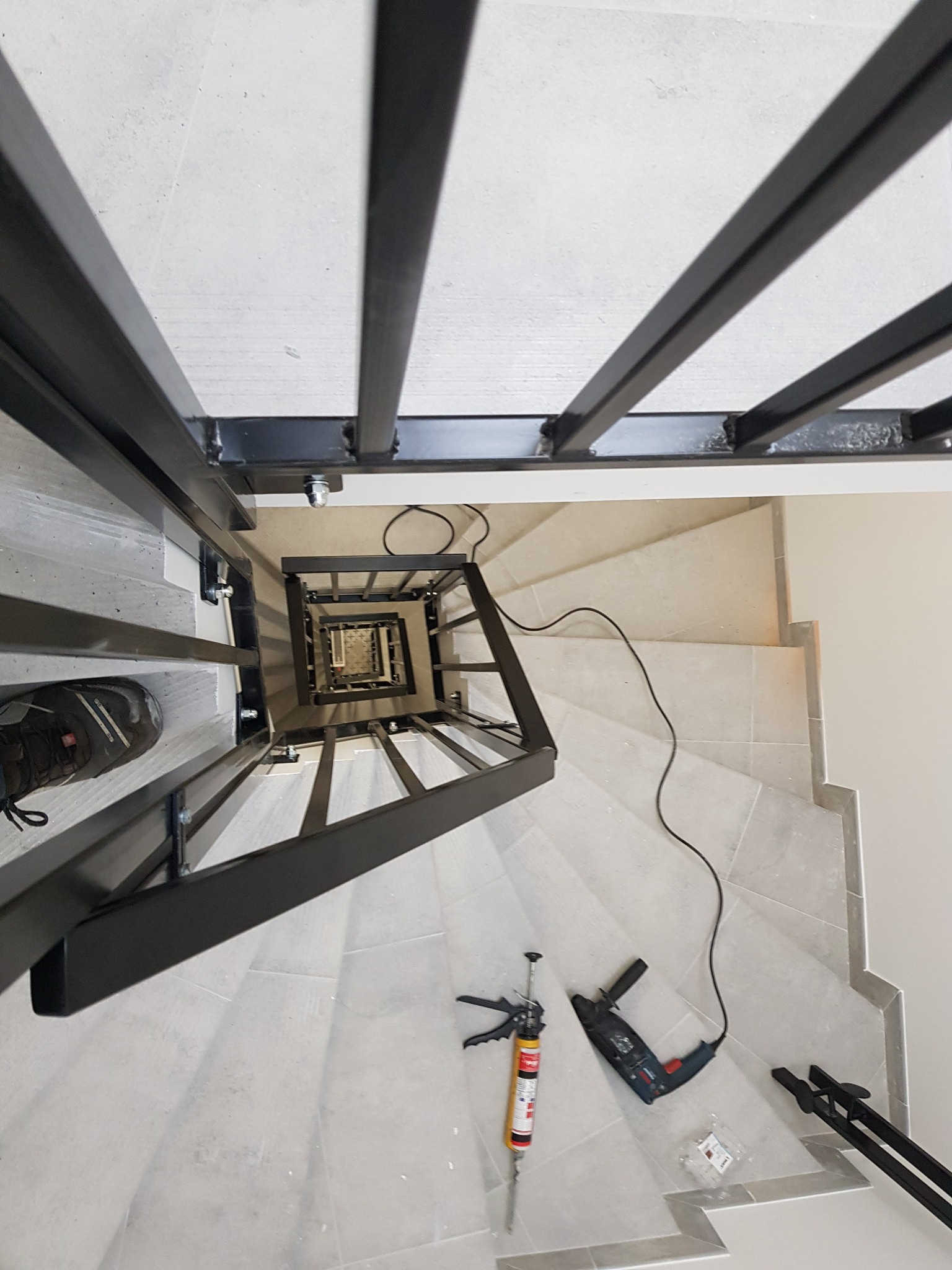 Widok z góry na spiralne schody z czarną metalową konstrukcją, w trakcie montażu, widoczne narzędzia: wiertarka, pistolet do silikonu, oraz stopa w bucie roboczym.