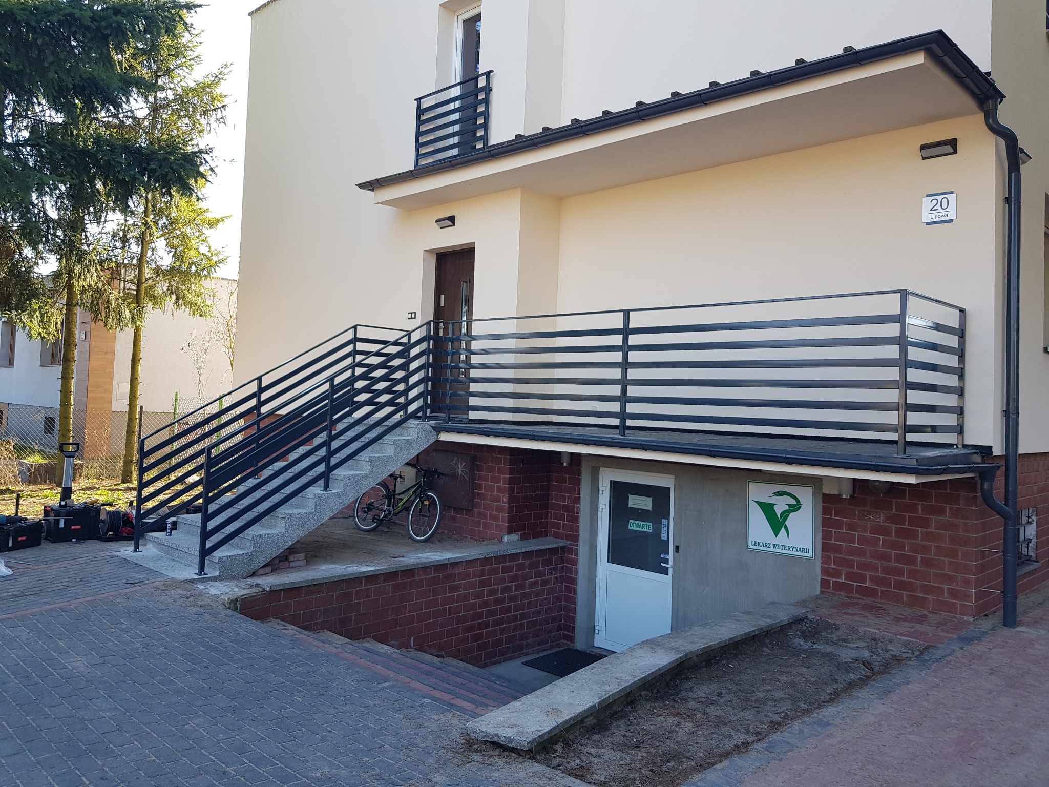Zewnętrzne schody z szarymi stopniami i czarną, poziomą barierką, prowadzące do drzwi wejściowych budynku z jasną elewacją; obok wejścia do gabinetu weterynaryjnego.