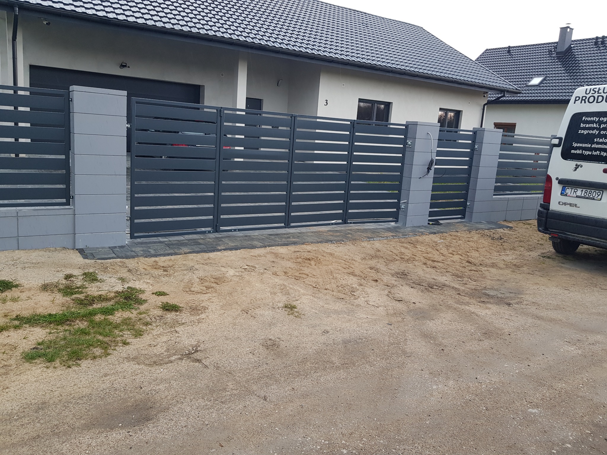 Nowoczesne, poziome ogrodzenie panelowe w kolorze grafitowym z szarymi słupkami betonowymi, oddzielające posesję z białym domem od piaszczystego terenu. Częściowo widoczny samochód dostawczy...