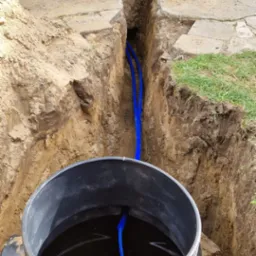 Montaż studni głębinowej: czarny zbiornik z niebieskimi rurami w wykopie ziemnym, widoczne wejście do budynku w tle.