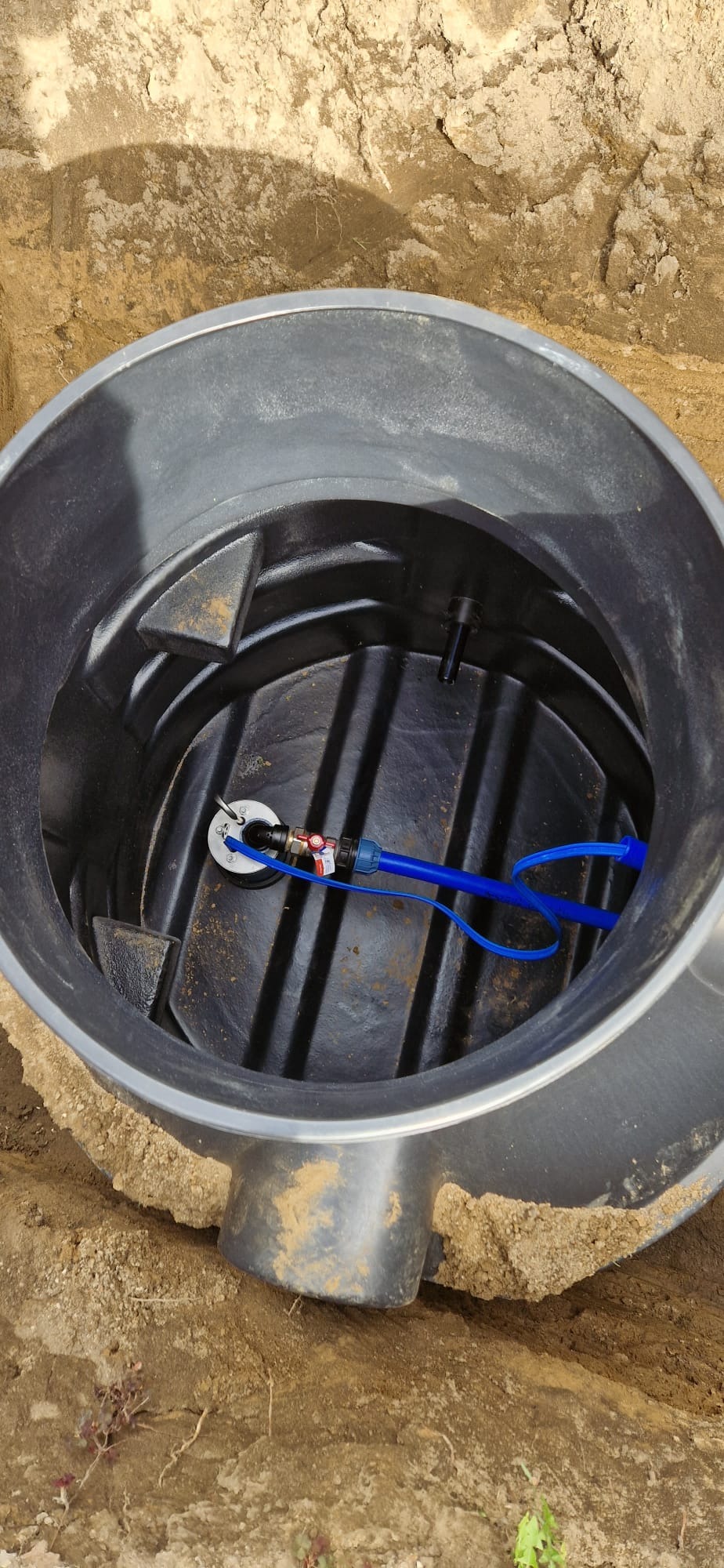 Wnętrze studni z czarnego tworzywa sztucznego, widoczne przyłącze z zaworem i niebieską rurką, całość umieszczona w wykopie z ziemi.
