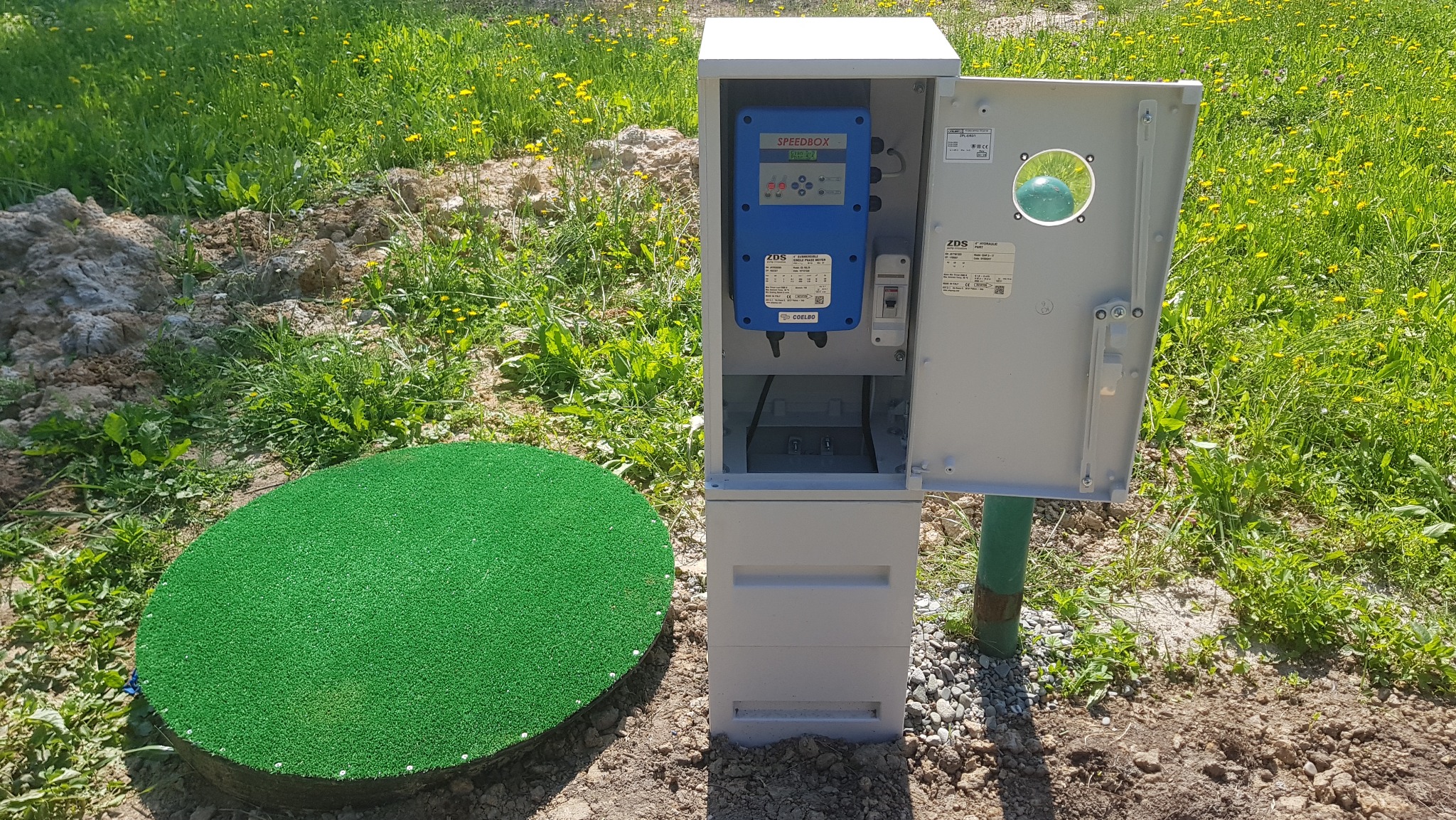 Zewnętrzna instalacja z szarą szafką sterowniczą z otwartymi drzwiczkami, niebieskim panelem sterowania SPEEDBOX i zieloną pokrywą studni na trawiastym terenie.