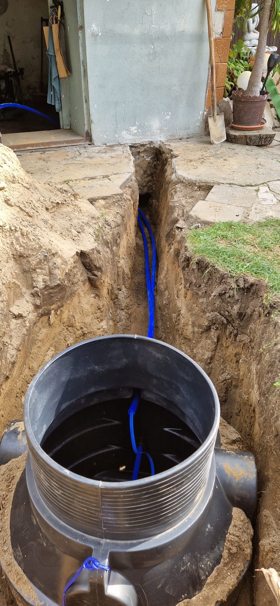 Montaż studni głębinowej: czarny zbiornik z niebieskimi rurami w wykopie ziemnym, widoczne wejście do budynku w tle.