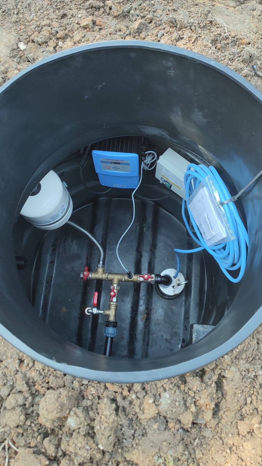 Wnętrze czarnej studzienki z zainstalowaną armaturą, pompą i sterownikiem Speedbox, widok z góry.
