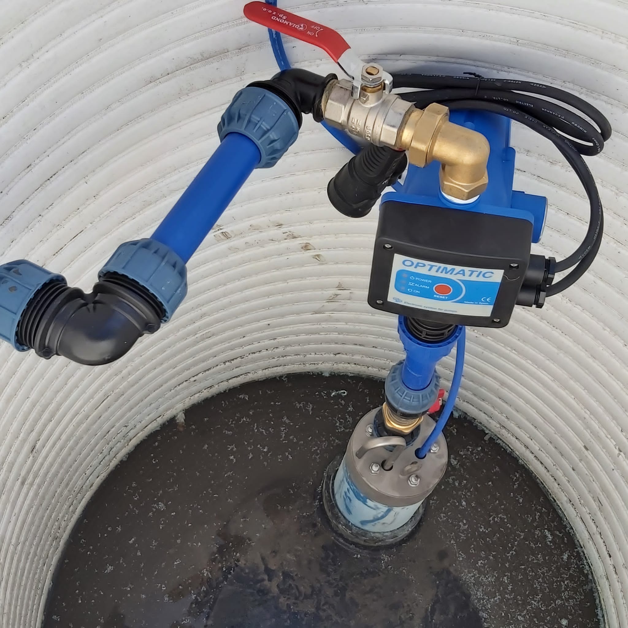 Pompa zanurzona w studni z widocznymi rurami i zaworem, zamontowana wewnątrz białej, karbowanej obudowy studni.