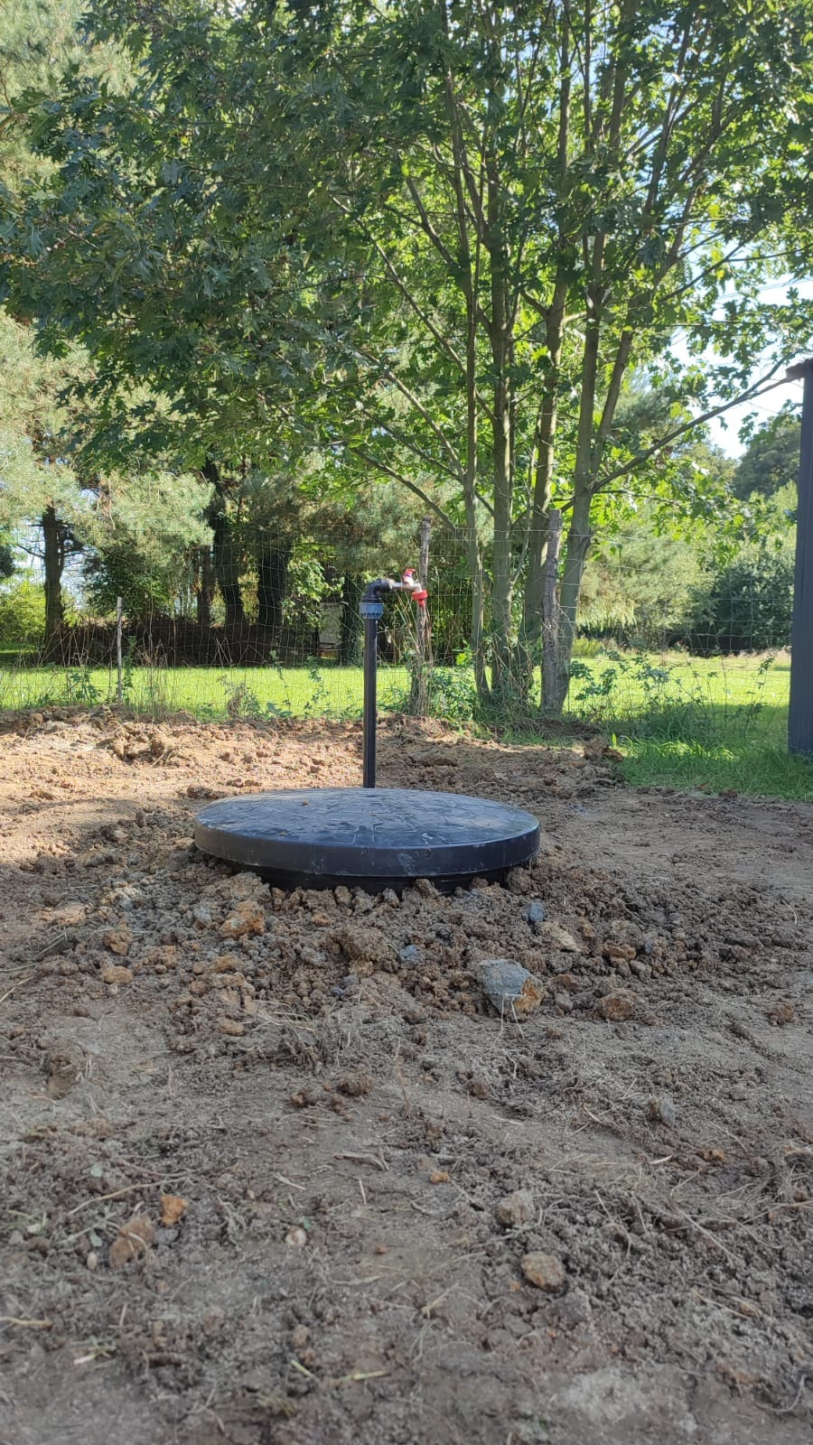 Zakończona instalacja studni z czarną pokrywą w ziemi, z metalową rurą i kranem na tle drzew i ogrodzenia.