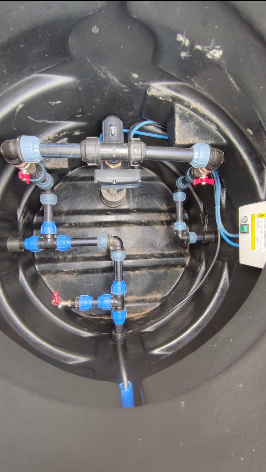 Wnętrze czarnego zbiornika z instalacją wodną: rury z niebieskimi złączkami, zawory z czerwonymi kurkami oraz licznik przepływu wody. Widoczna również skrzynka elektryczna z przełącznikiem.